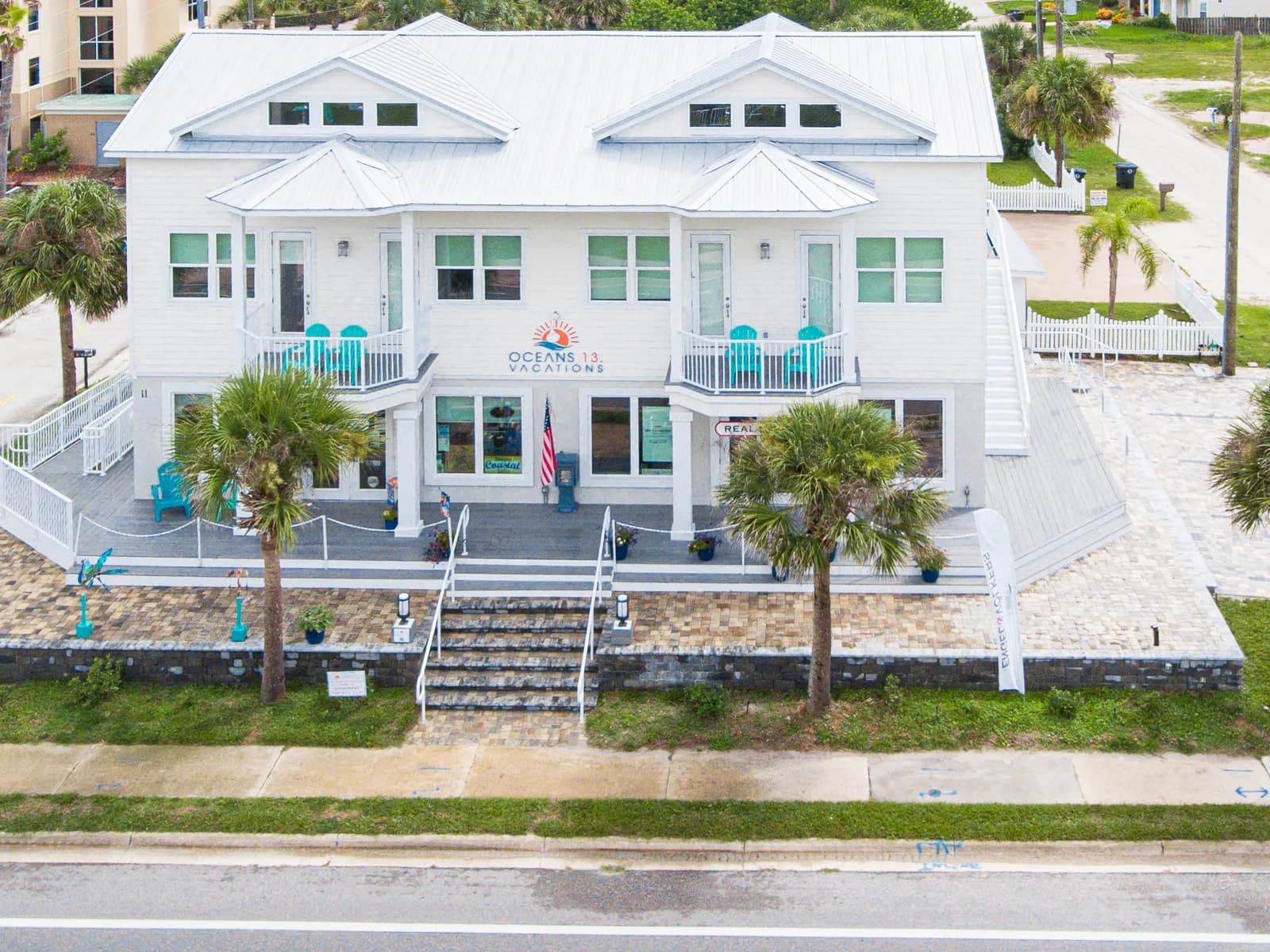 The Engel & Völkers St. Augustine shop on 14th Lane in St. Augustine Beach, a white two-storey building with a paved courtyard and palms, the Atlantic behind it