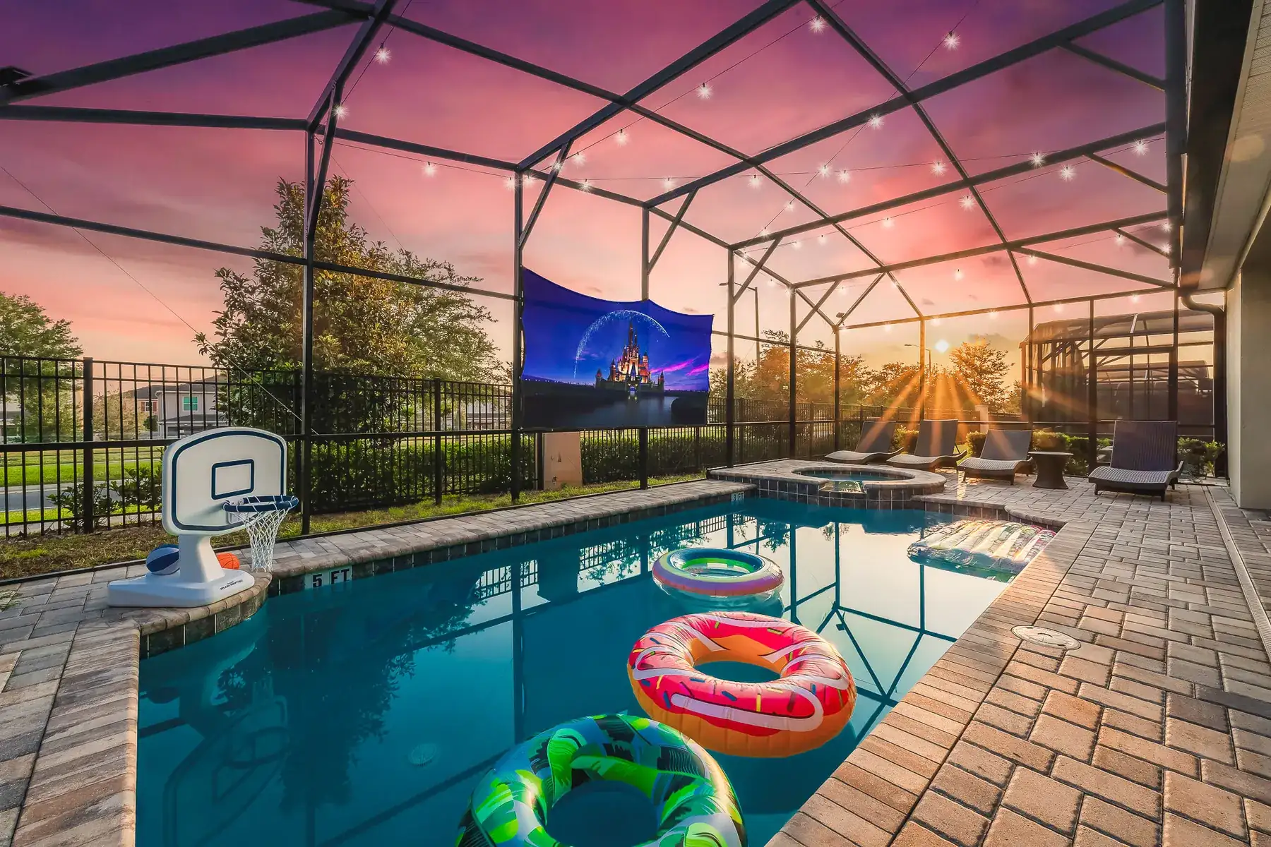Screened pool at sunset with an outdoor movie screen showing a castle and fireworks, string lights overhead and floats on the water.