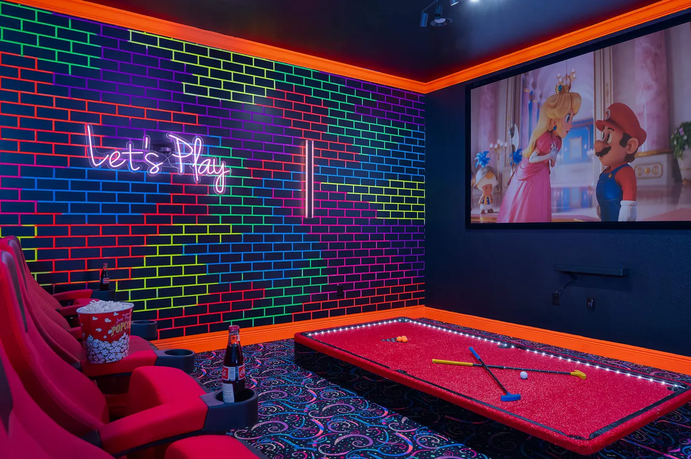 Home theater and game room lit in neon, with red reclining theater seats, a red-felt pool table and a wall-size screen.